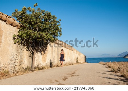 Young woman with sunglasses walking next to a wall with a tree in an empty road in a sunny day. Mediterranean sea in the background.