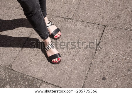 Model wearing black and white sandals with zebra print. Red nail varnish on toes and skinny black jeans. Focus on woman legs and feet. Shot from above, pavement in the background.
