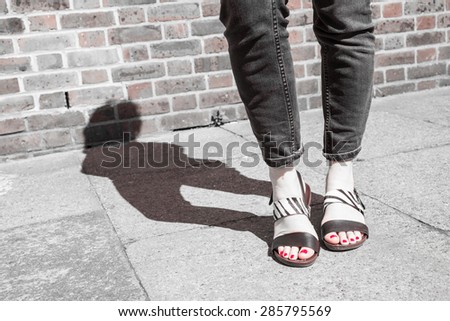 Model wearing black and white sandals with zebra print. Red nail varnish on toes and skinny black jeans. Focus on woman legs and feet. Shadow and brick wall in background. Black and white.