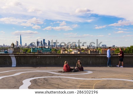 Peckham, London, UK - June 06 2015: Two girls and two guys drinking beer on the floor of a rooftop bar cafe in south-east London.View of the City of London skyscrapers and the Shard in the background.