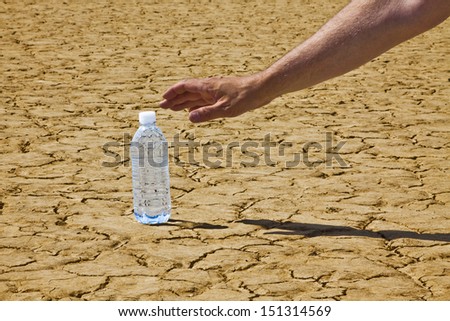 A man\'s hand and arm is reaching in from the upper right corner down to a bottle of water sitting on a desert playa.