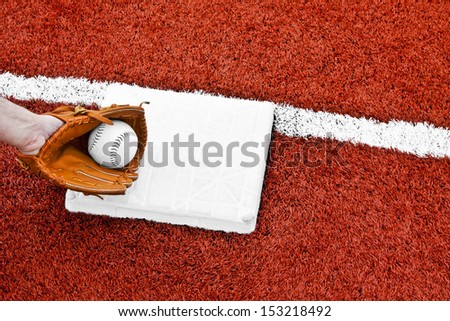 The hand in the mitt has the softball and tagging the base to put the runner out before reaching the base in the middle of the red artificial turf.