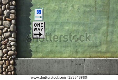 The handicap symbol arrow sign points in the same direction as the one way sign. Signs are mounted on a stucco wall above the foundation and next to rock column. Large area for text copy on the right.