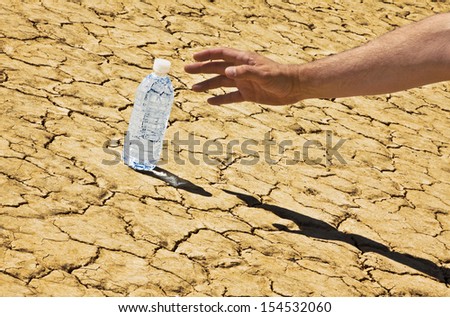A manÃ¢Â?Â?s hand and arm is reaching in from the right side down to a bottle of water sitting on a desert playa. The whole image is tilted.