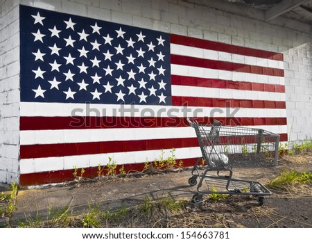 The old shopping cart is sitting on a sidewalk, with an American flag painted on a concrete block wall behind it. Consider at the current time that American is in bad shape and portrayed by the photo.