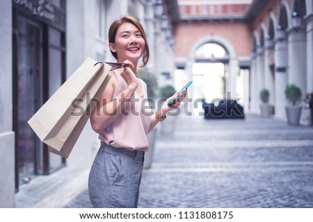 Beautiful Asian Thai girl in shopping.Happy woman with shopping, girls holding shopping bags using a smart phone and smiling while standing outdoors enjoying in shopping.Consumerism,lifestyle concept.