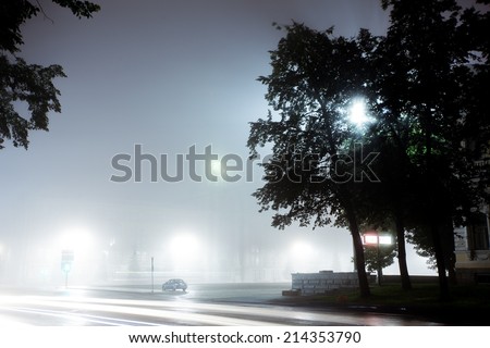 A lonely car drives along empty city street at night after rain. Street is filled with neon light blurred with fog