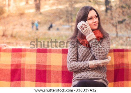 Young beautiful woman in a knitted sweater scarf and gloves sitting on a bench covered with a rug against autumn park background