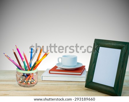 Photo frame, books, coffee cup, color pencils on wooden table over grey wall background/ interior still life