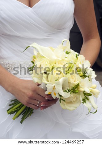 Close-up of white roses and freesias wedding bouquet in a hands of a slim bride in a white wedding dress.