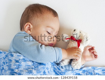 Portrait of a little baby boy sleeping under a blue blanket holding a white soft toy teddy bear.