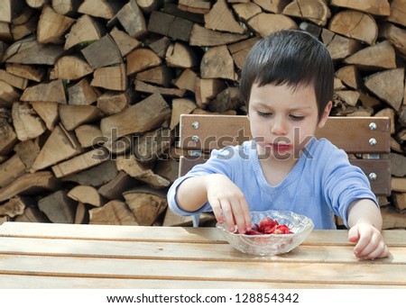 Child eating strawberries at the outside wooden table on a patio or at a garden.