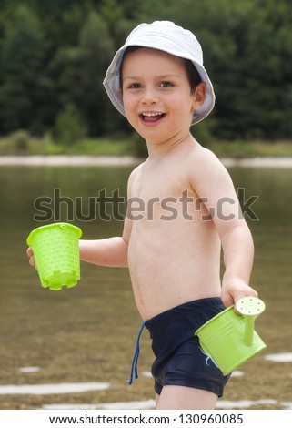 Portrait of a little boy playing with watering can and plastic toy in a river or lake.