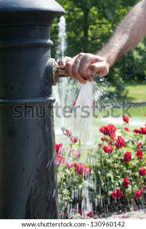 A  hand on a tap of a  water fountain with running water and a rose garden or a park in the background.