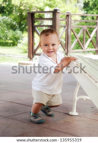 Happy toddler child leaning against a bench in a spring park or a patio garden, trying to get up, learning to walk.