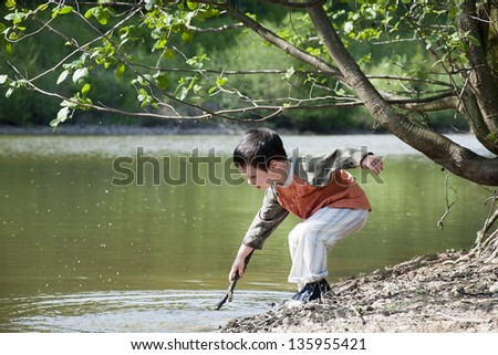 Child boy playing with a stick on the edge of the lake or a river in the nature.