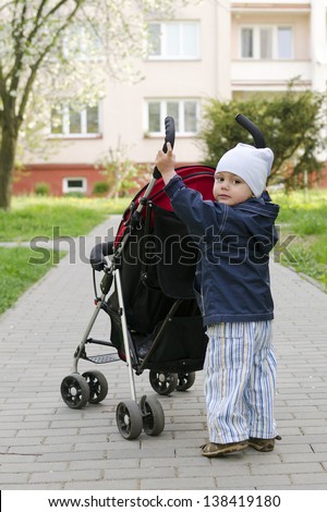 Small child toddler, boy or girl, pushing a stroller on a pedestrian path.