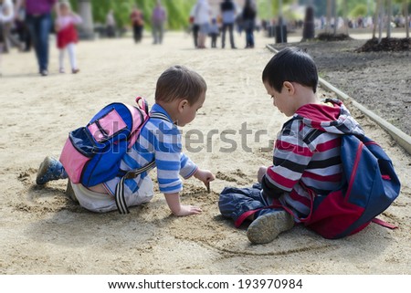 Two children sitting on ground drawing into sand path with stick.