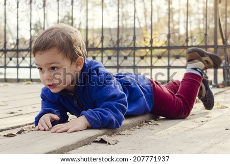 Child boy laying on a wooden floor ground on a bridge or landing in autumn or fall park outside.