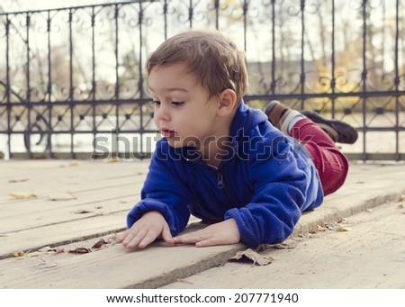 Child boy laying on a wooden floor ground on a bridge or landing in autumn or fall park outside.