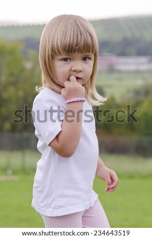 Portrait of a cut child girl with a finger in her mouth biting her nail standing in park or garden in summer.
