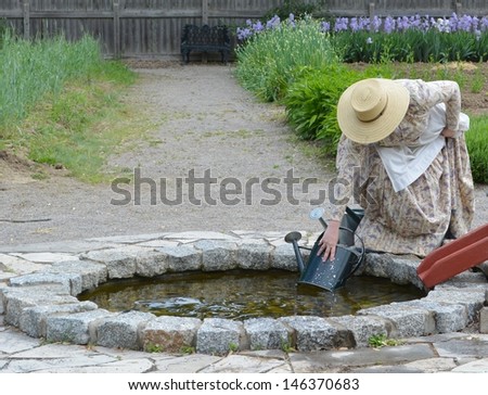 unrecognizable female in turn of century clothing bending down and filling a watering can at a well in a garden