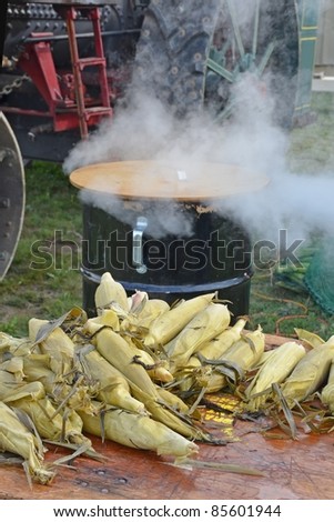 steamed corn on the cobs laying on a wooden table, steaming kettle with emerging steam in the background