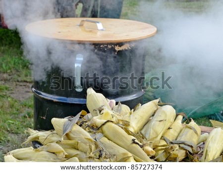 steamed corn on the cobs laying on a wooden table, steaming kettle with emerging steam in the background