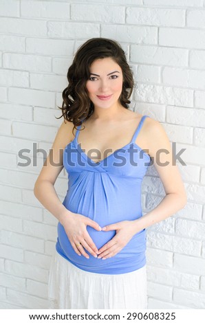 young beautiful pregnant woman in blue shirt with heart from hands on her belly on white brick wall background