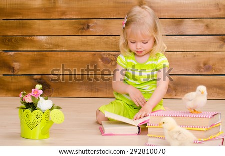 little blonde girl reading books and alive chickens on brown wooden wall background