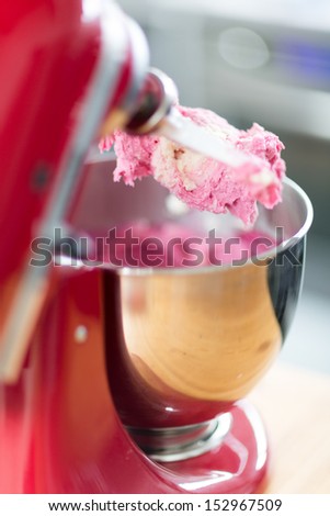 Close up view from behind of an electric mixer in a kitchen with colorful cake mix adhering to the beater which is raised above a stainless steel mixing bowl