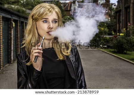 Pretty blond woman smoking an e-cigarette standing in a street in a leather jacket exhaling a cloud a smoke from her mouth while looking off to the side