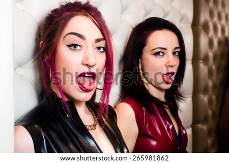 Close up Two Stylish Young Female Rock Stars Showing Tongue Piercing While Leaning on the Wall and Looking at the Camera.
