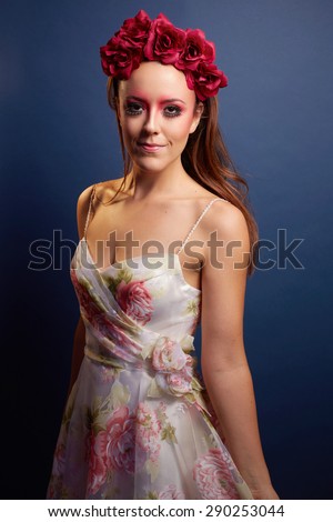 Three quarter length close up of serene, smiling 20s brunette woman with heavy makeup wearing floral dress and large scarlet flowers in hair, on navy background