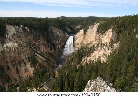 View of Lower Yellowstone Falls with Grand Canyon of the Yellowstone in Yellowstone National Park, with rough rock and trees on both sides. Blue sky with some clouds.