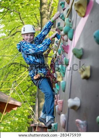 Teenager in helmet and with a safety rope climbing wall holding hooks on the blurred background of trees