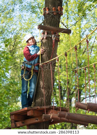 Adult woman in a helmet and with a safety system standing beside a tree and the suspended rope bridge on a blurred background
