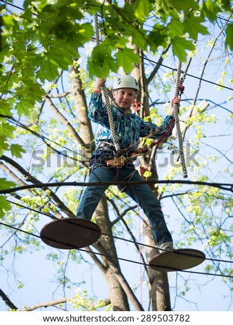 Little boy in the helmet and a safety system stands on the suspension bridge and keeps the ropes against the sky and the trees with leaves