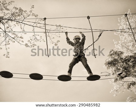 Toned image Teenager in the helmet and a safety system stands on the suspension bridge and keeps the ropes against the sky and the trees with leaves