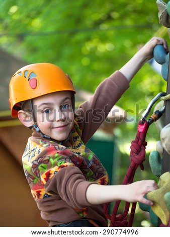 Teenager in helmet and with a safety rope climbing wall holding hooks on the blurred background of trees