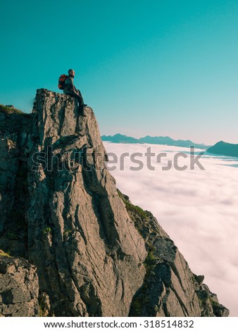 Toned image adult man with backpack sitting, legs dangling on the edge of a cliff and looks into the distance against the blue sky and thick clouds floating down