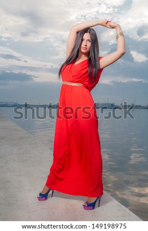 Fashion portrait of classy brunette in red shiny dress and long straight hair . vertical shot against sky, outdoors. luxury photography.