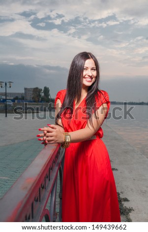 Smiling portrait of classy brunette in red shiny dress and long straight hair . Vertical shot against evening sky, outdoors. Luxury photography.