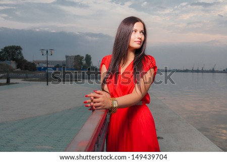 Fashion portrait of classy brunette looking away in red shiny dress and long straight hair . vertical shot against sky, outdoors. luxury photography.
