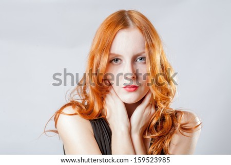 Horizontal beauty shot of a young blue eyed woman with her red hair, touching face by her hands, looks very shy