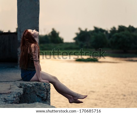 Red haired female looking up profile view. Redhead women sitting near river in summertime and dreaming. Sunset light
