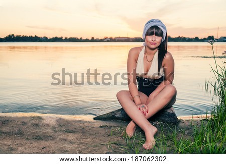 Vintage photo of relaxing young woman in nature. Retro fashion female sitting on lake shore and looking at camera