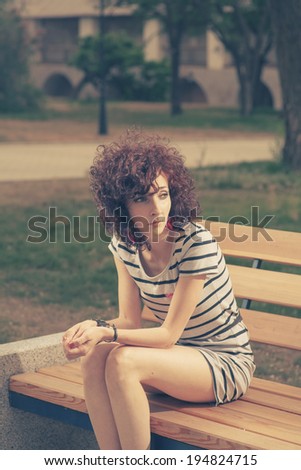 Summer portrait of a beautiful girl with curly red hair sitting in calm state on park bench and looking away