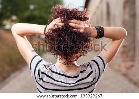 Spacious rear view of a girl with red curly hair tilting her head back and looking up to the sky near ancient ruins, outdoors.