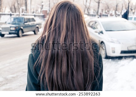 Rear view of a young woman covered with her long blond hair and looking at the street with cars.